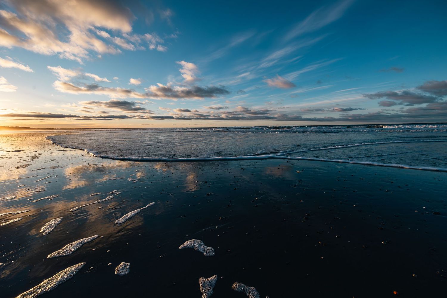 Dramatic sunset sky reflects on wet beach sand with dark water and clouds creating mirror-like effect at golden hour.