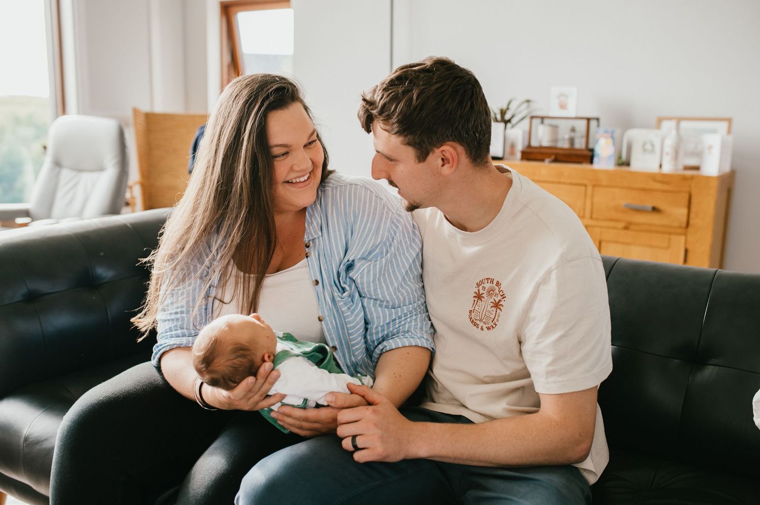 A couple sits together on a dark couch while cradling their newborn baby in a bright, cozy living room setting.