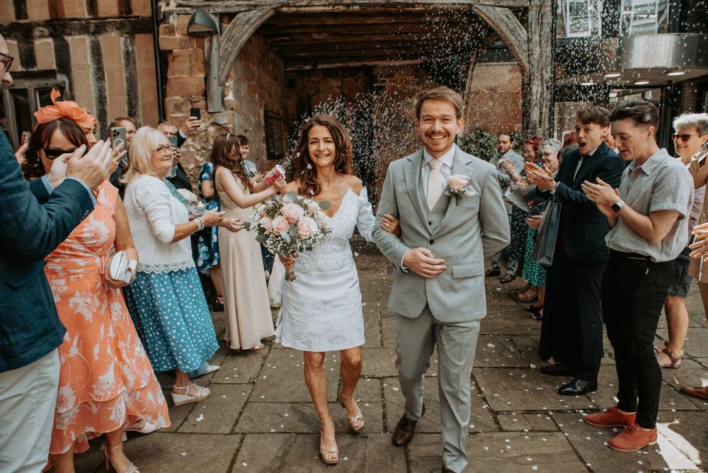 Wedding guests shower newlyweds with confetti as they exit a rustic brick archway during outdoor celebration.