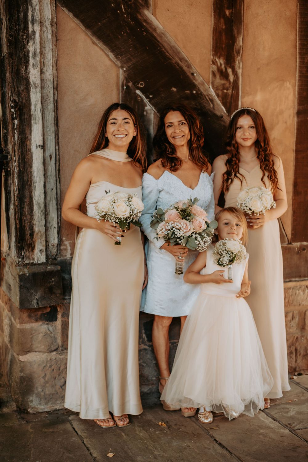 Wedding party members in cream and light blue dresses pose together holding floral bouquets at a rustic venue.