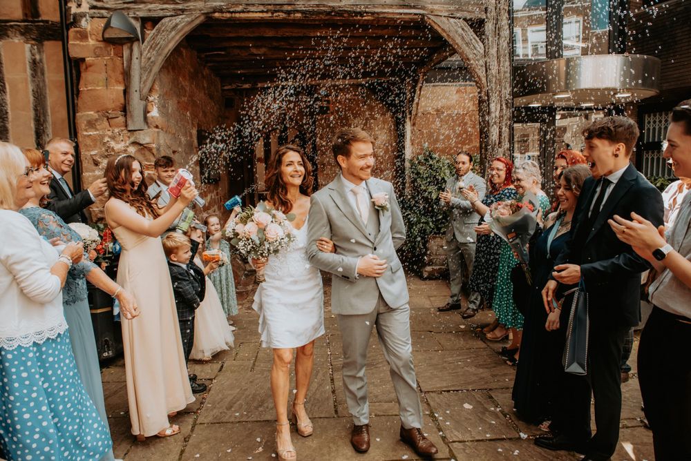 Wedding guests throw confetti on newlyweds exiting stone archway during joyful outdoor ceremony celebration.