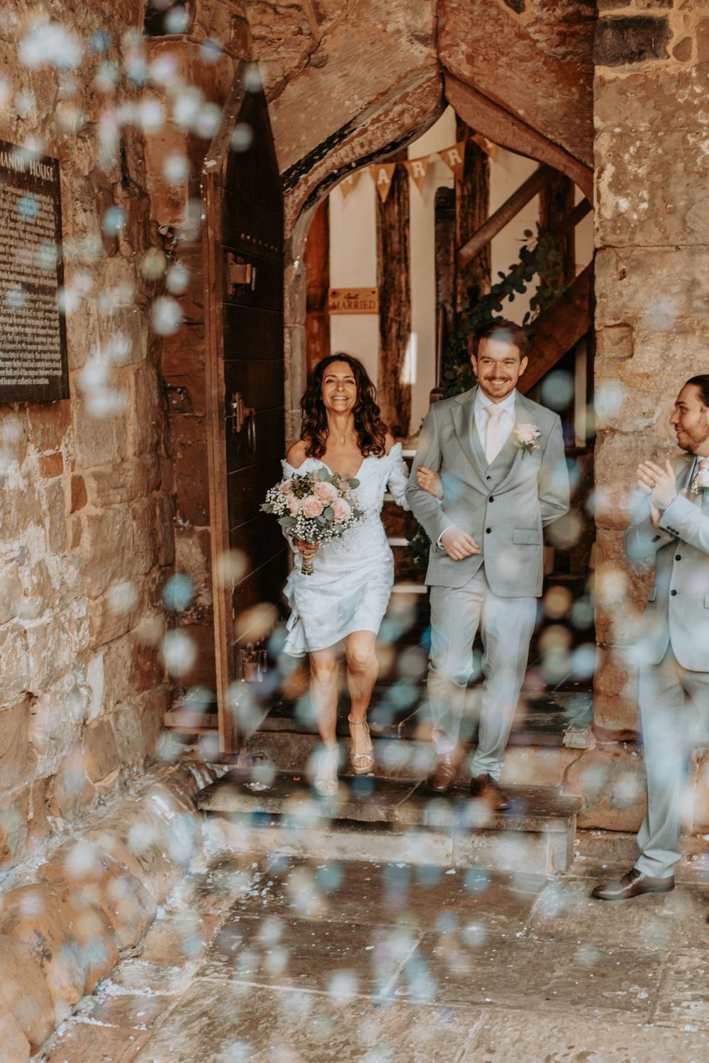 A wedding couple joyfully exits a rustic stone church while bokeh light effects dance across the foreground.