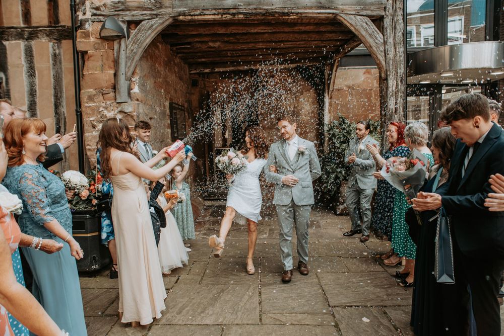 Wedding guests toss confetti over a newlywed couple exiting through a rustic stone archway at an outdoor ceremony.