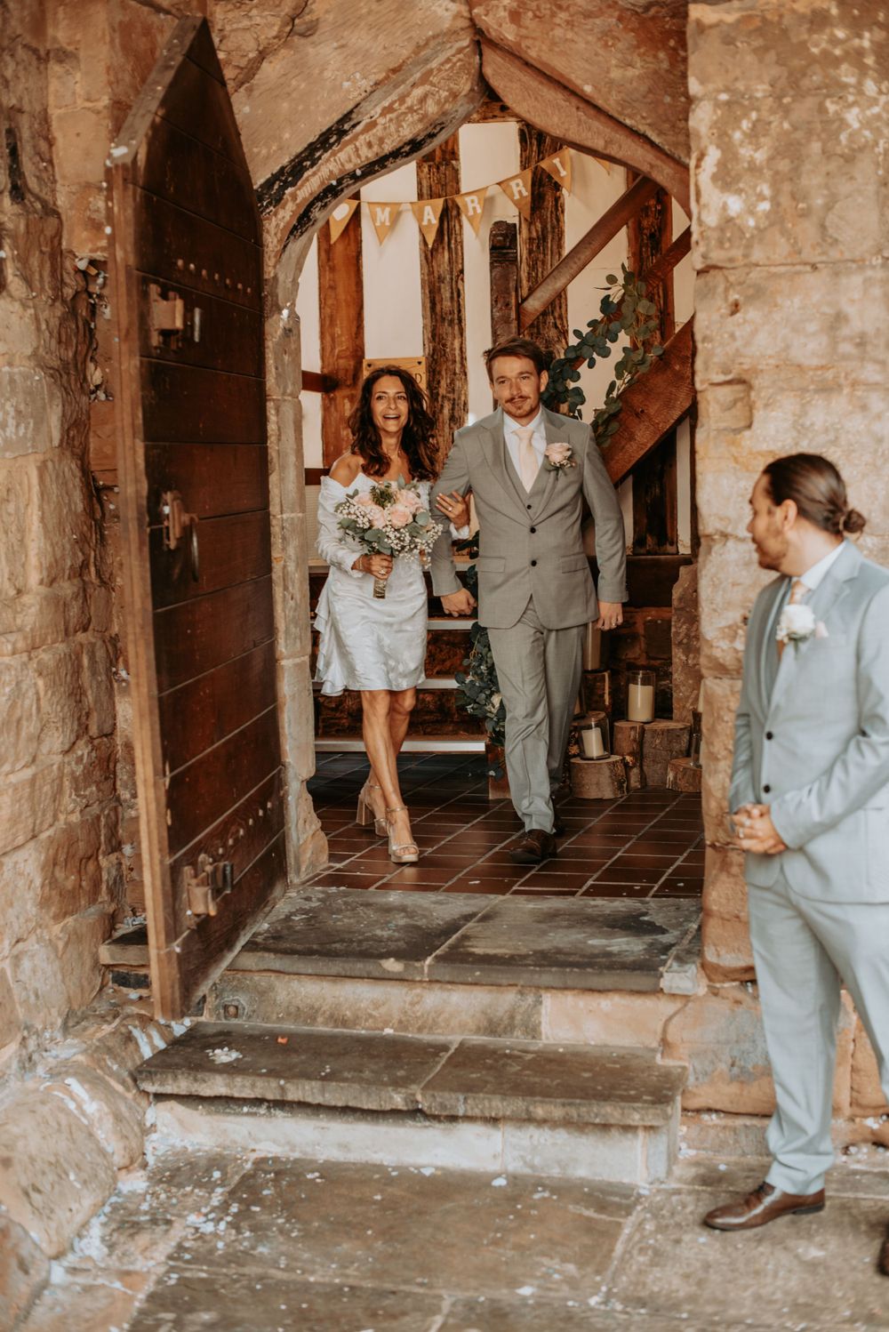 Couple exits rustic stone church doorway after their intimate wedding ceremony in vintage boho style.