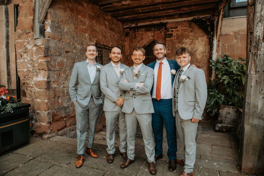 Wedding party of five groomsmen wearing gray suits and one in blue posing against rustic brick wall at venue.