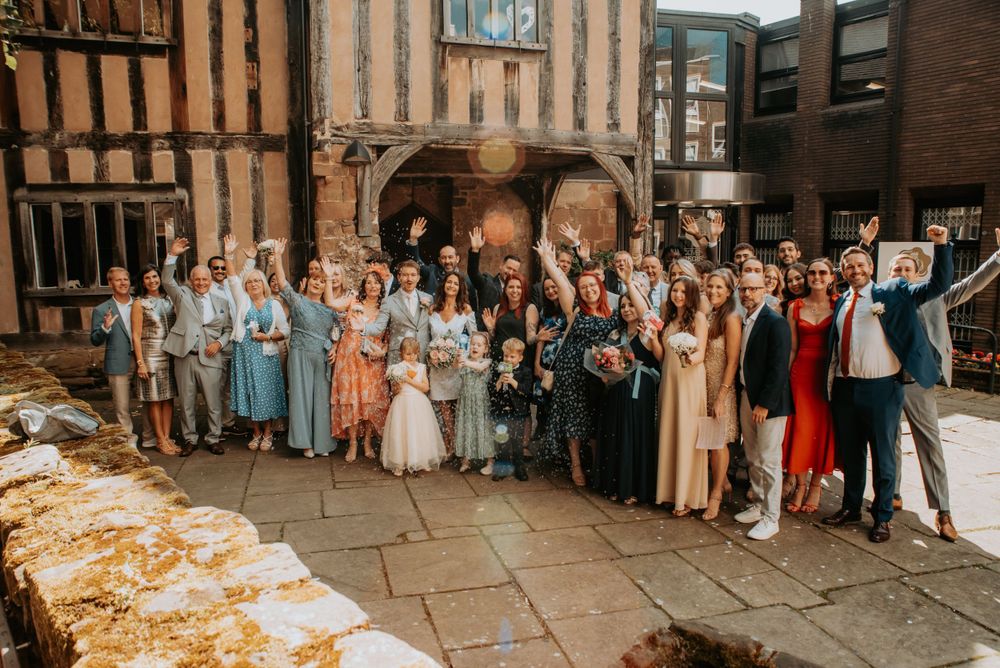 A large wedding party poses together outside a historic brick building with vintage architectural details.