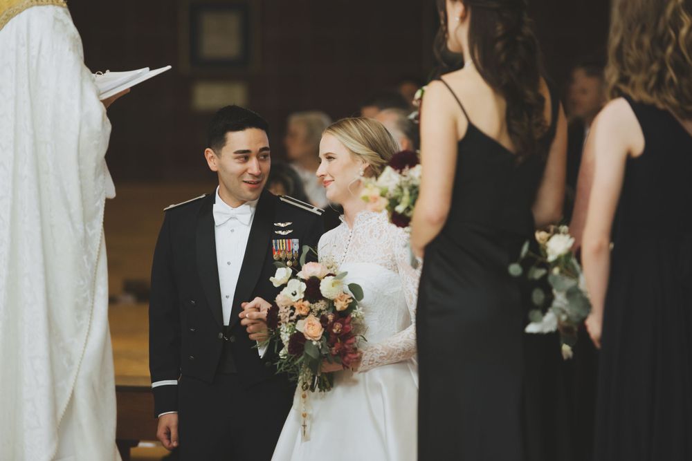 Wedding ceremony with bride in white dress holding floral bouquet standing next to groom alongside bridesmaids in black.