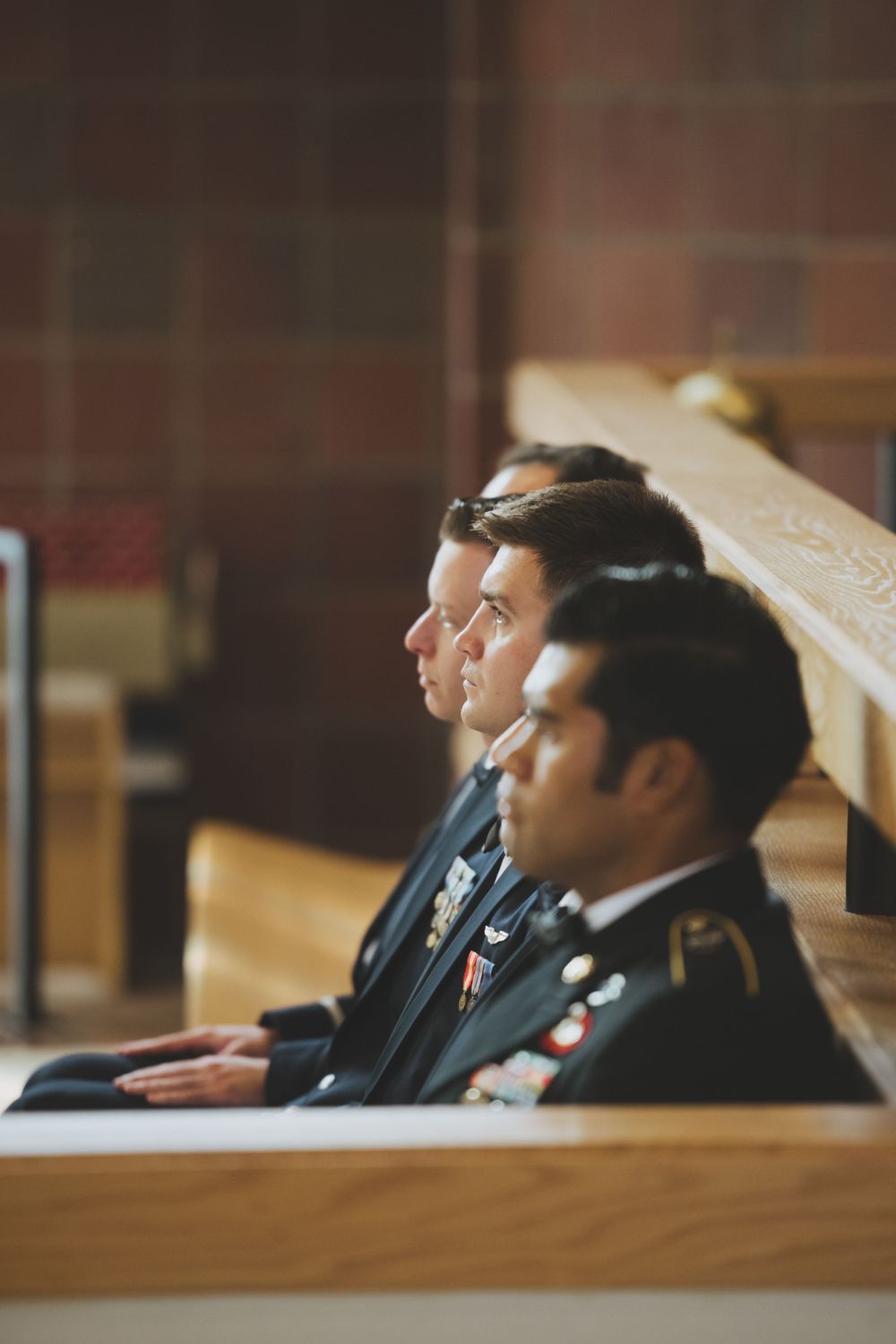 Groomsmen in formal suits sit together in a wooden church pew during a wedding ceremony.