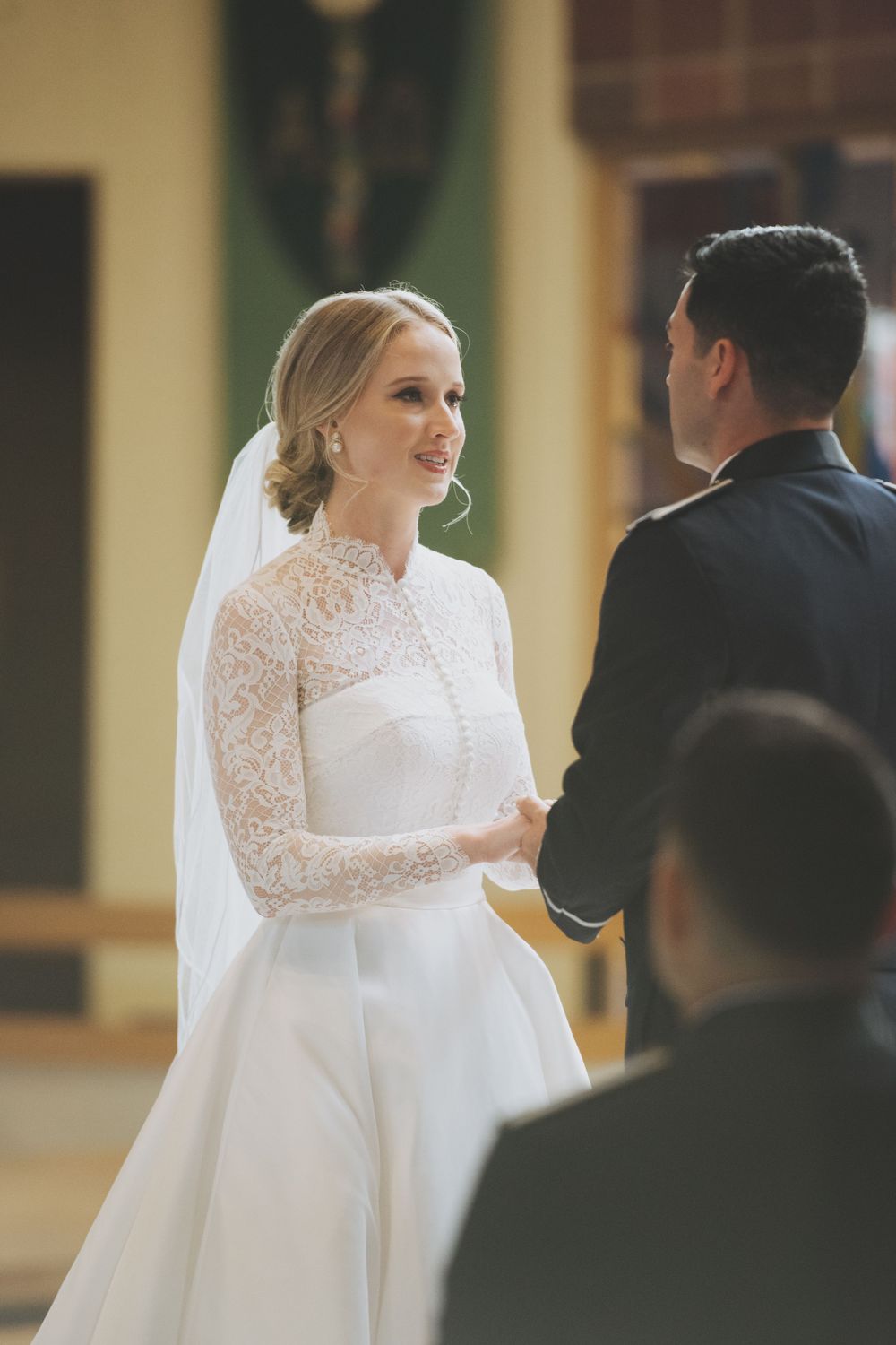 A bride in a long-sleeved lace wedding dress and veil faces groom during a traditional ceremony.