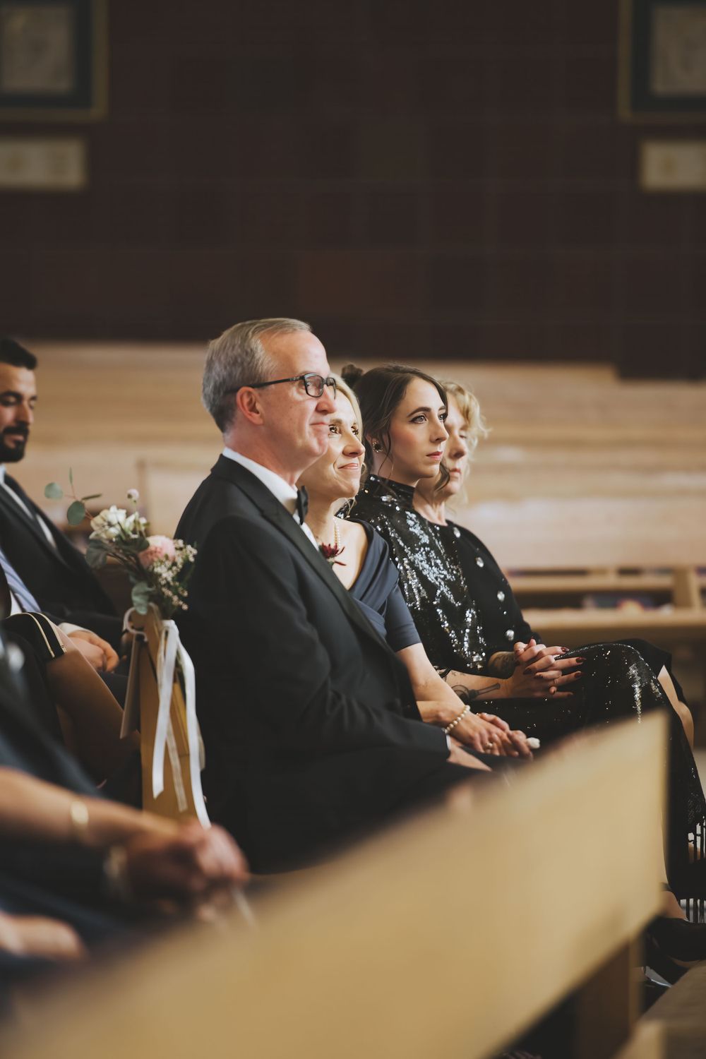 People sitting in wooden church pews during a wedding ceremony, with formal attire and floral decorations visible.