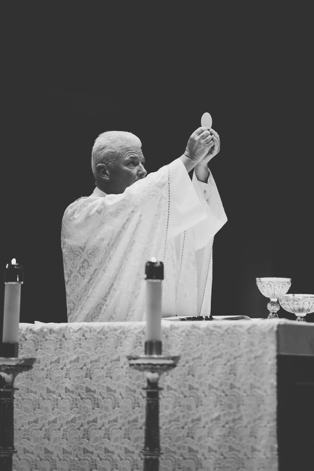 Catholic priest raises communion host during mass at altar with candles in black and white photograph.