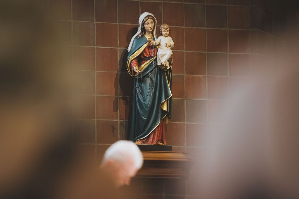 Religious statue depicting Madonna and child mounted on a brown tiled wall in a church setting.
