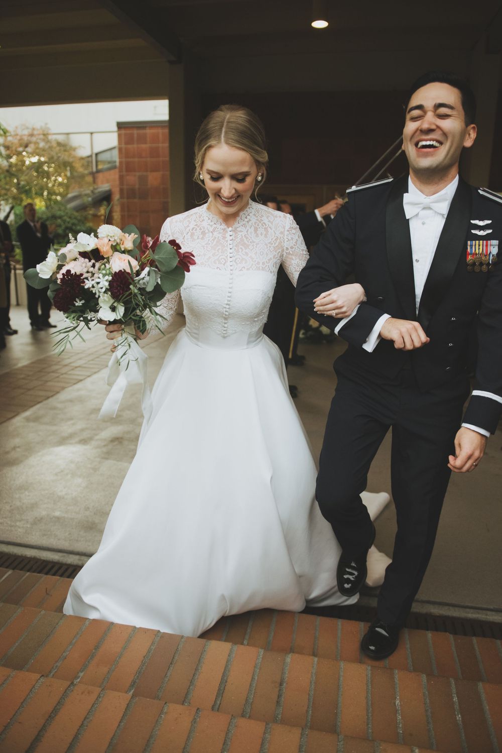 A joyful wedding couple exits their venue, with the bride in a lace white gown and groom in military formal dress uniform.