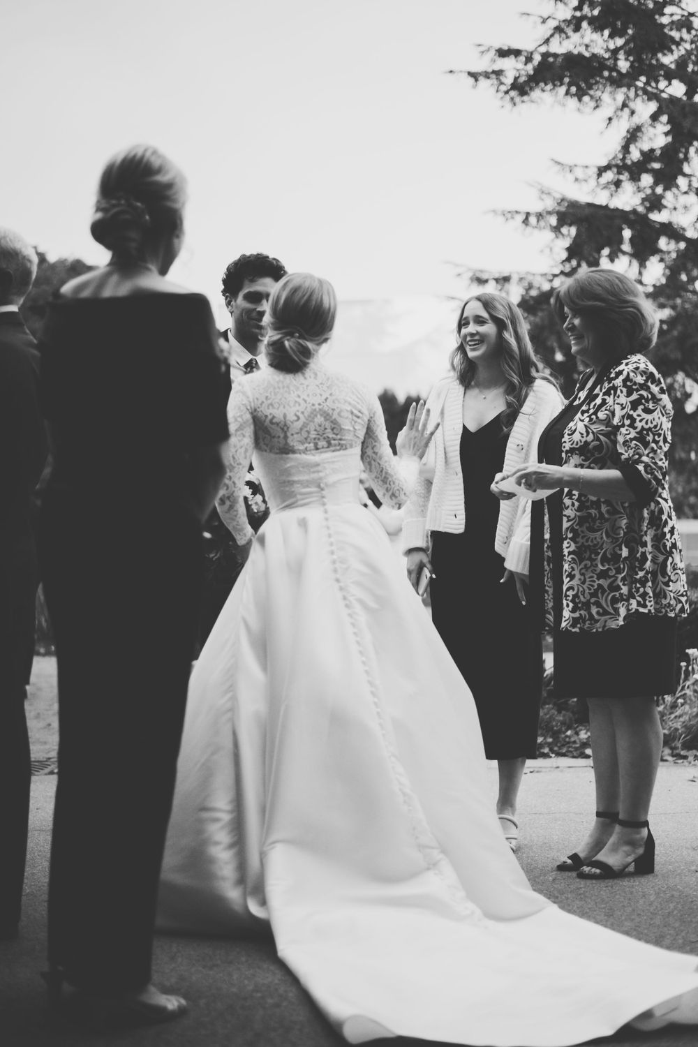 A black and white wedding photo showing a bride in a flowing lace dress surrounded by guests at an outdoor reception.