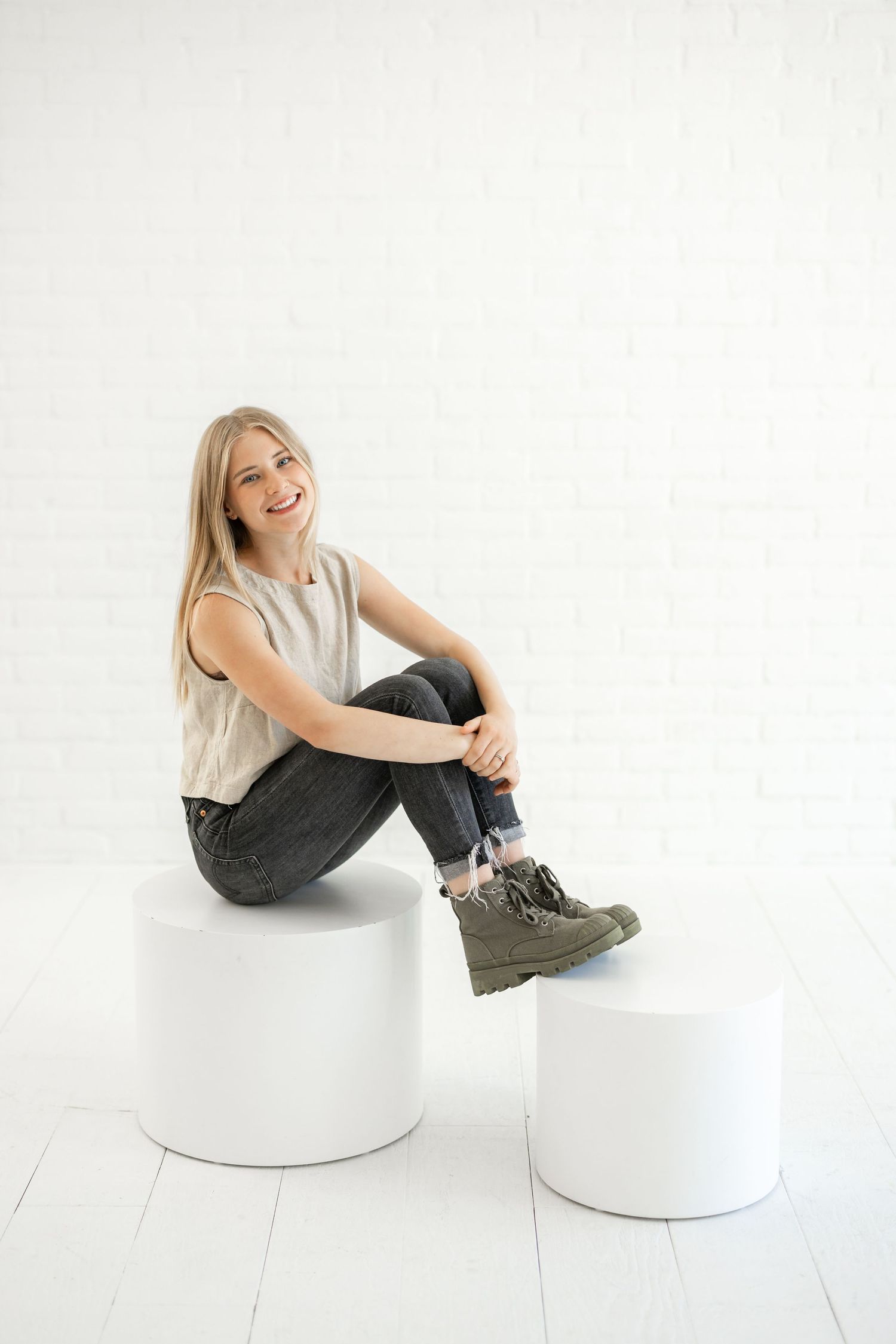 Woman in casual attire smiles while sitting on white cylindrical pedestal.