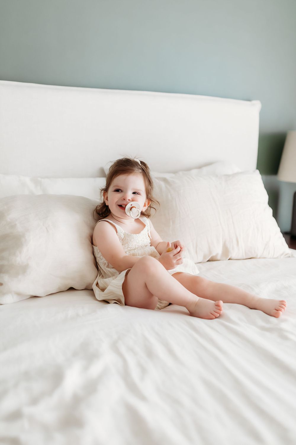 Small child in white clothing sits smiling on crisp white bedding against a pale blue wall.