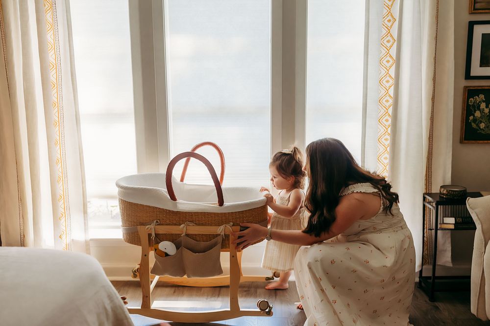 A tender moment beside a wooden rocking bassinet in a sunlit room with flowing curtains in the background.