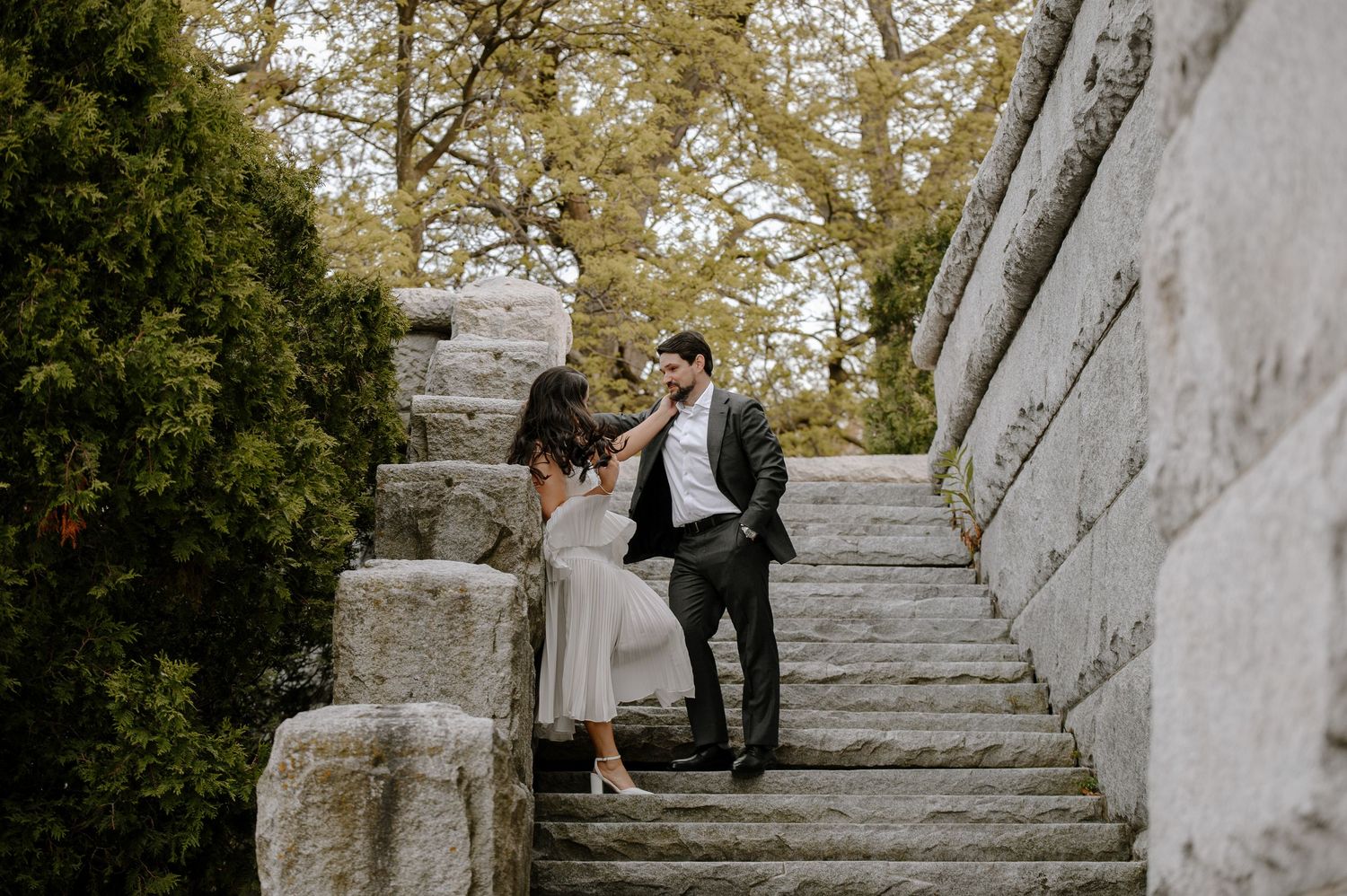 A romantic couple embraces on an elegant white stone staircase in an outdoor setting during a wedding photoshoot.