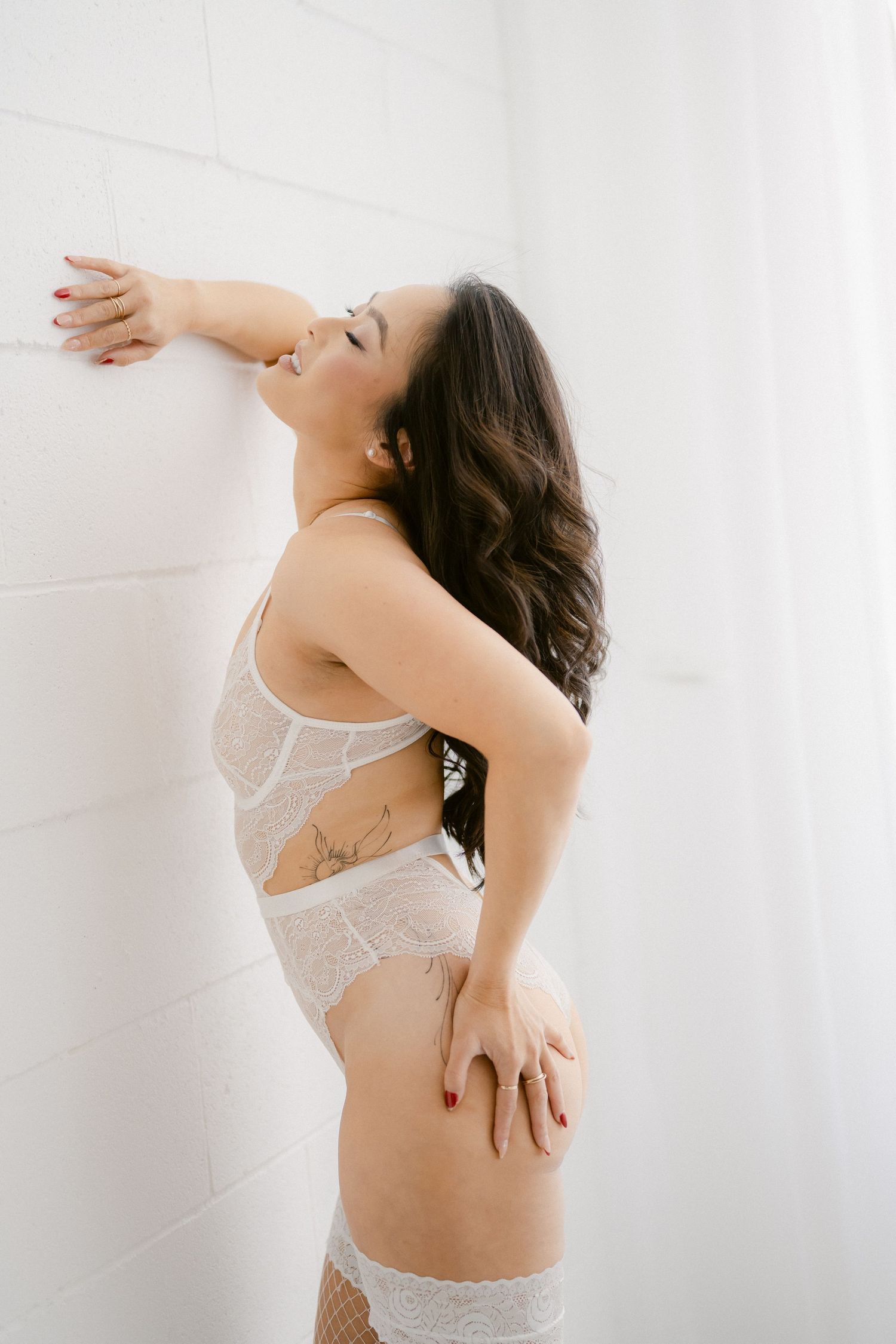 Woman in white lace lingerie poses against white wall in artistic boudoir photography session.