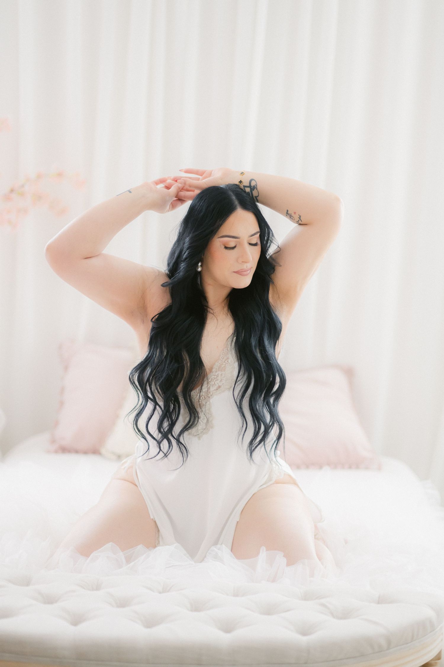 Dreamy boudoir photography featuring long dark wavy hair and white bodysuit on soft pink and white bedding.