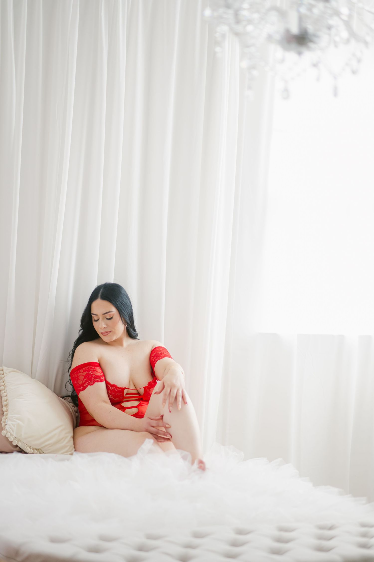 A person in red lingerie sits on a white bed next to sheer curtains in a softly lit boudoir setting.