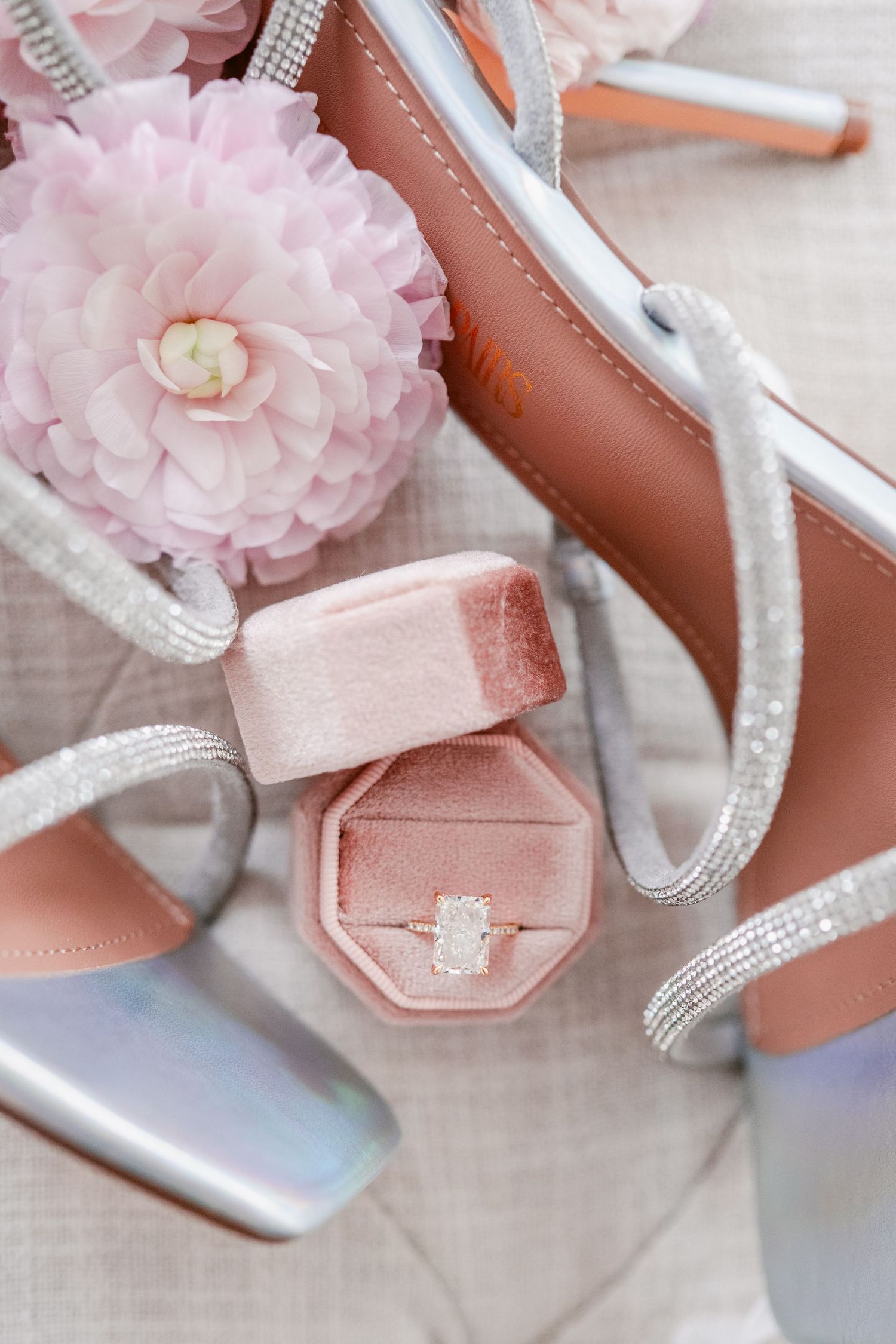 Pink engagement ring box with silver heels and blush flower on light fabric background.