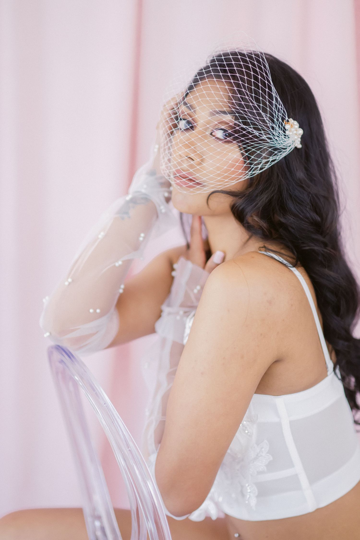 Elegant bridal boudoir photo featuring delicate white lingerie and vintage-style birdcage veil against pink backdrop.
