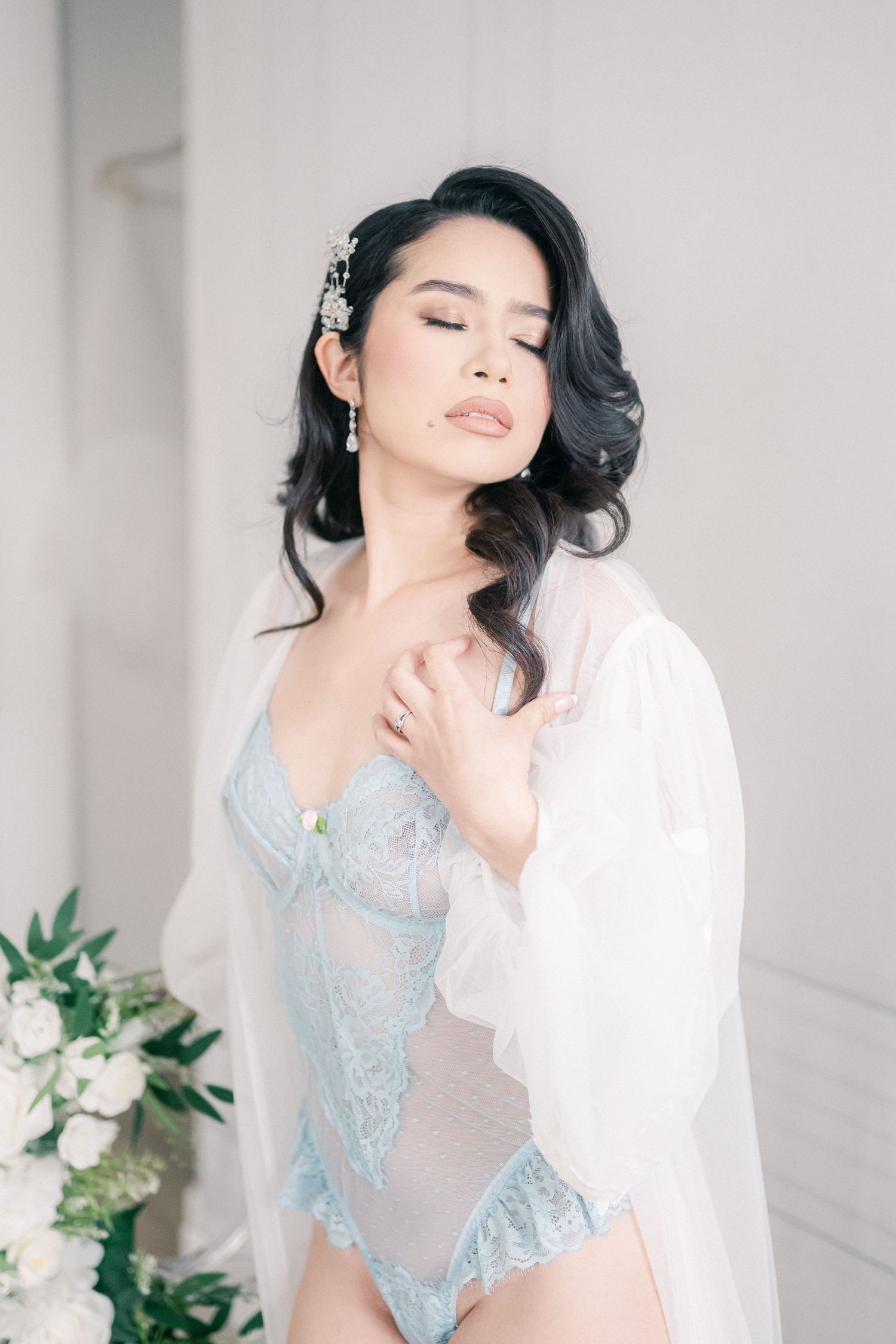 Romantic boudoir portrait with powder blue lace bodysuit and sheer white robe in minimalist setting.