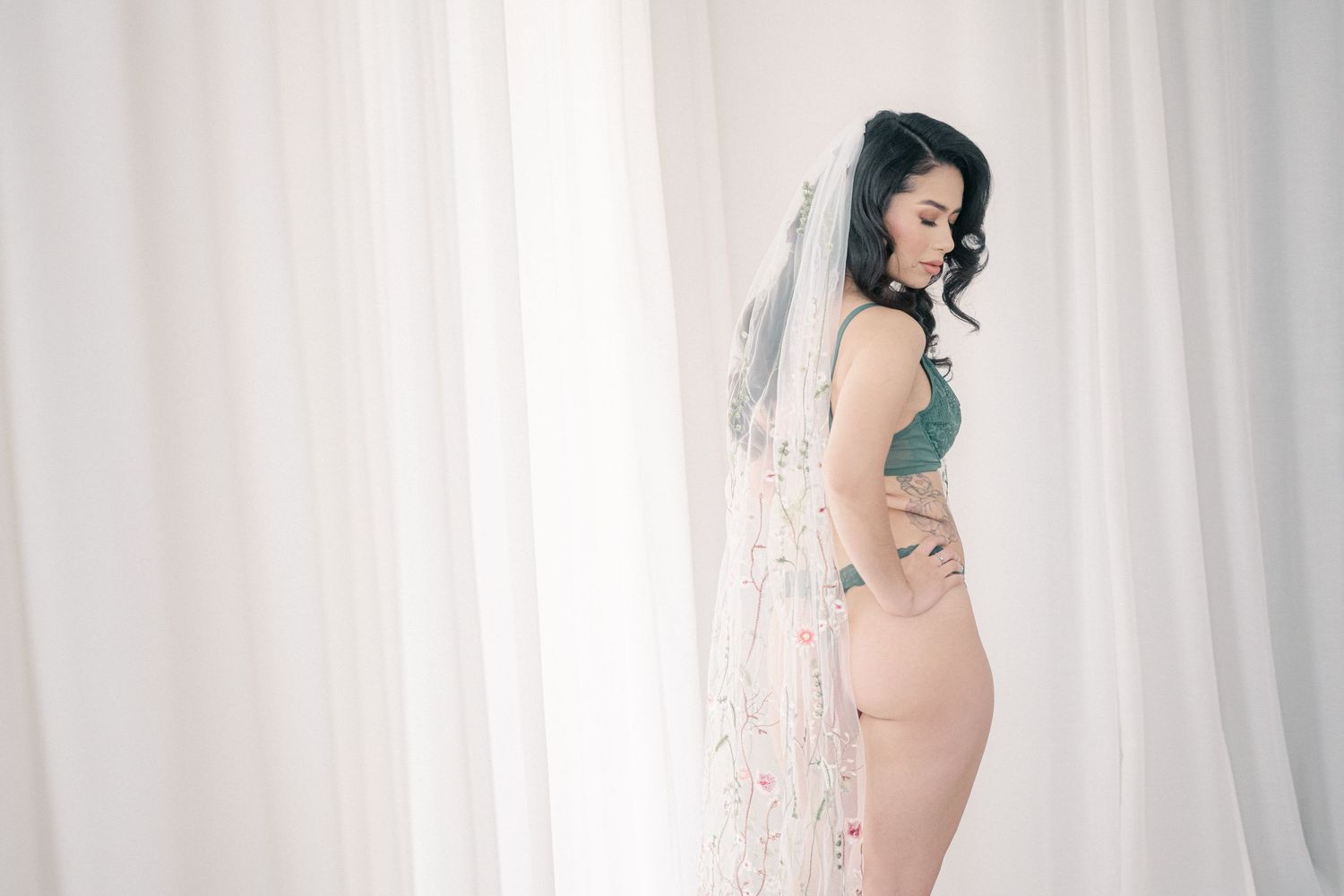 A figure poses elegantly in a white bridal veil against a light backdrop in a boudoir photography session.