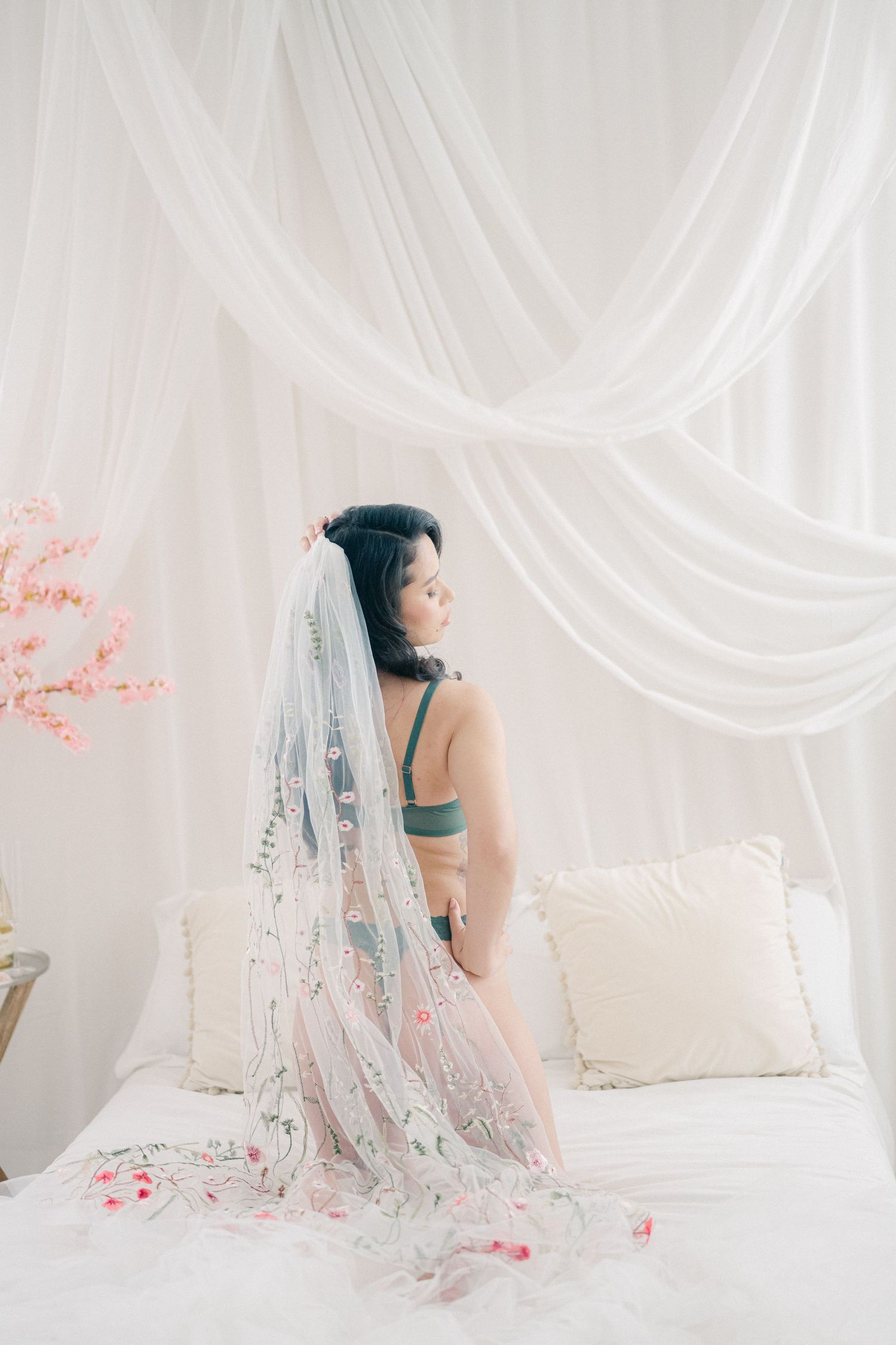 Artistic boudoir photo on white bed with flowing sheer veil adorned with pink floral details.