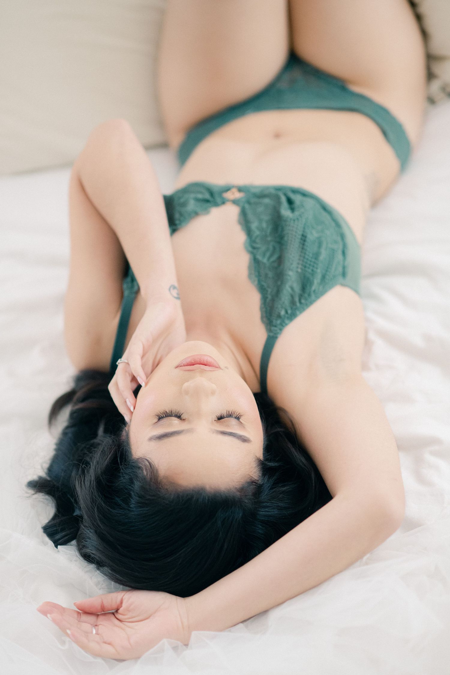 Person in teal lingerie lounging on white bedding in sensual boudoir photography pose.