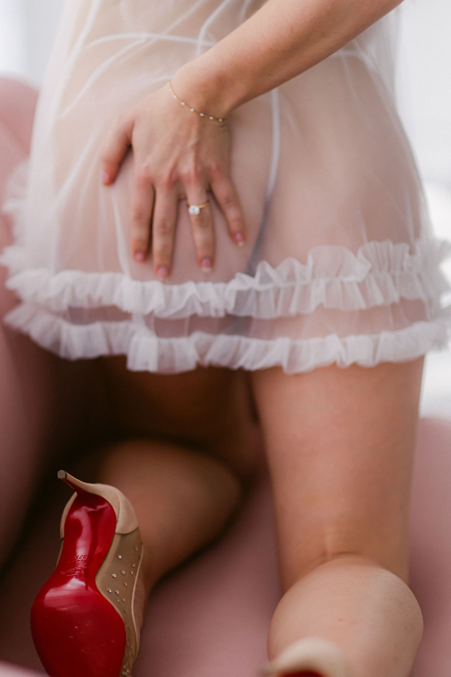 Close-up detail of a tulle skirt hem and red-soled designer shoe against a soft pink background.