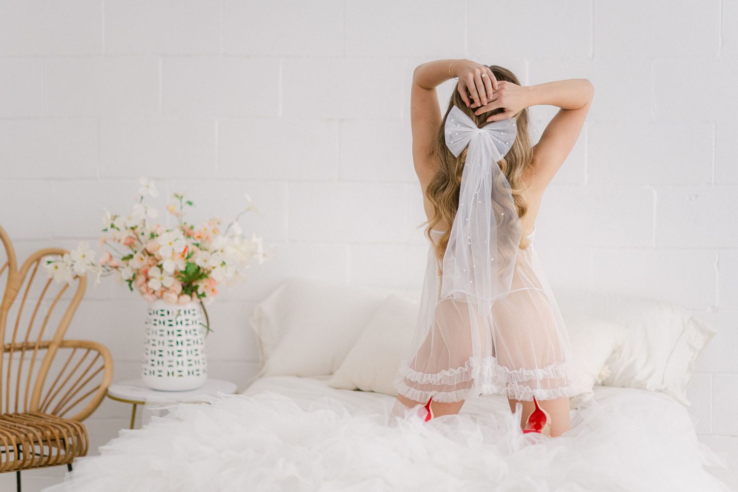 Dreamy portrait of someone in sheer robe standing on white bedding with floral arrangement in background.