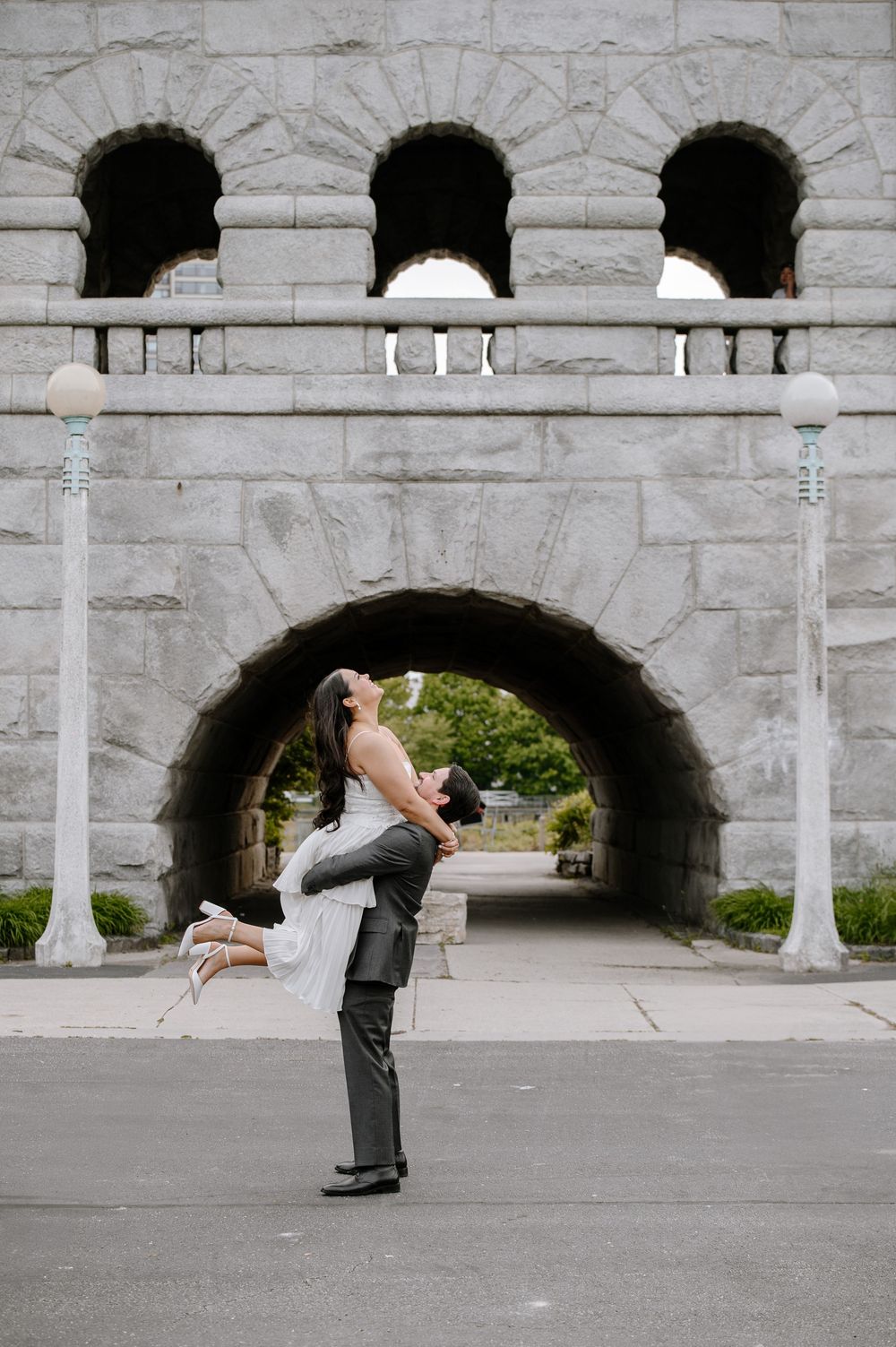 A romantic couple shares a joyful embrace under a stone archway with decorative architectural details.