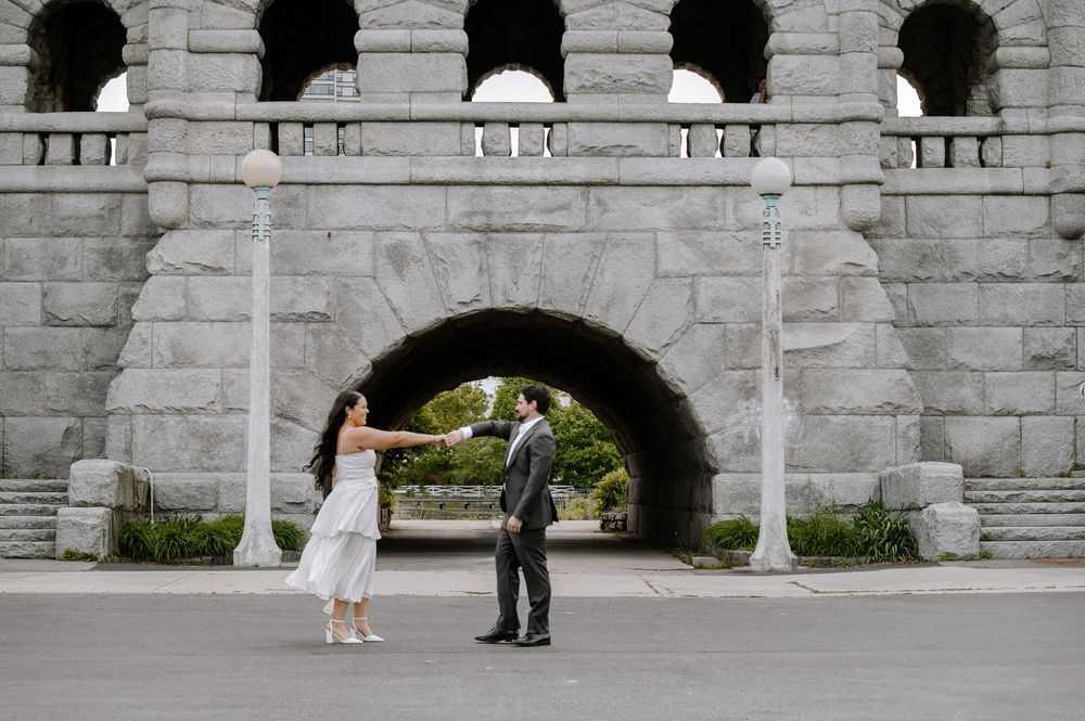 A couple holding hands and dancing in front of a stone archway with classic architectural details.