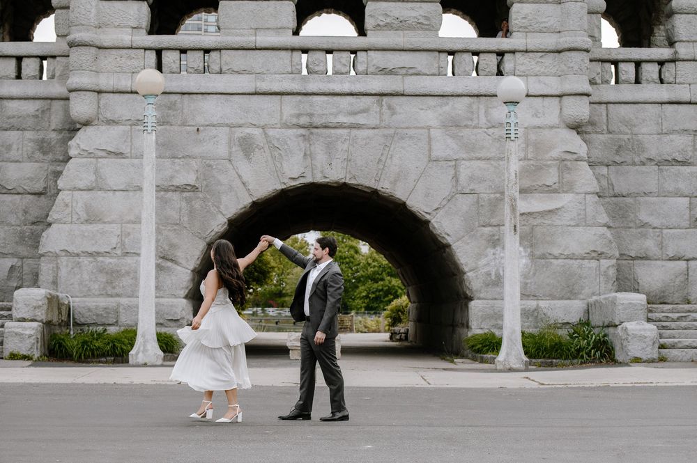 Two people in formal attire stand under a stone archway on a gray path surrounded by greenery.
