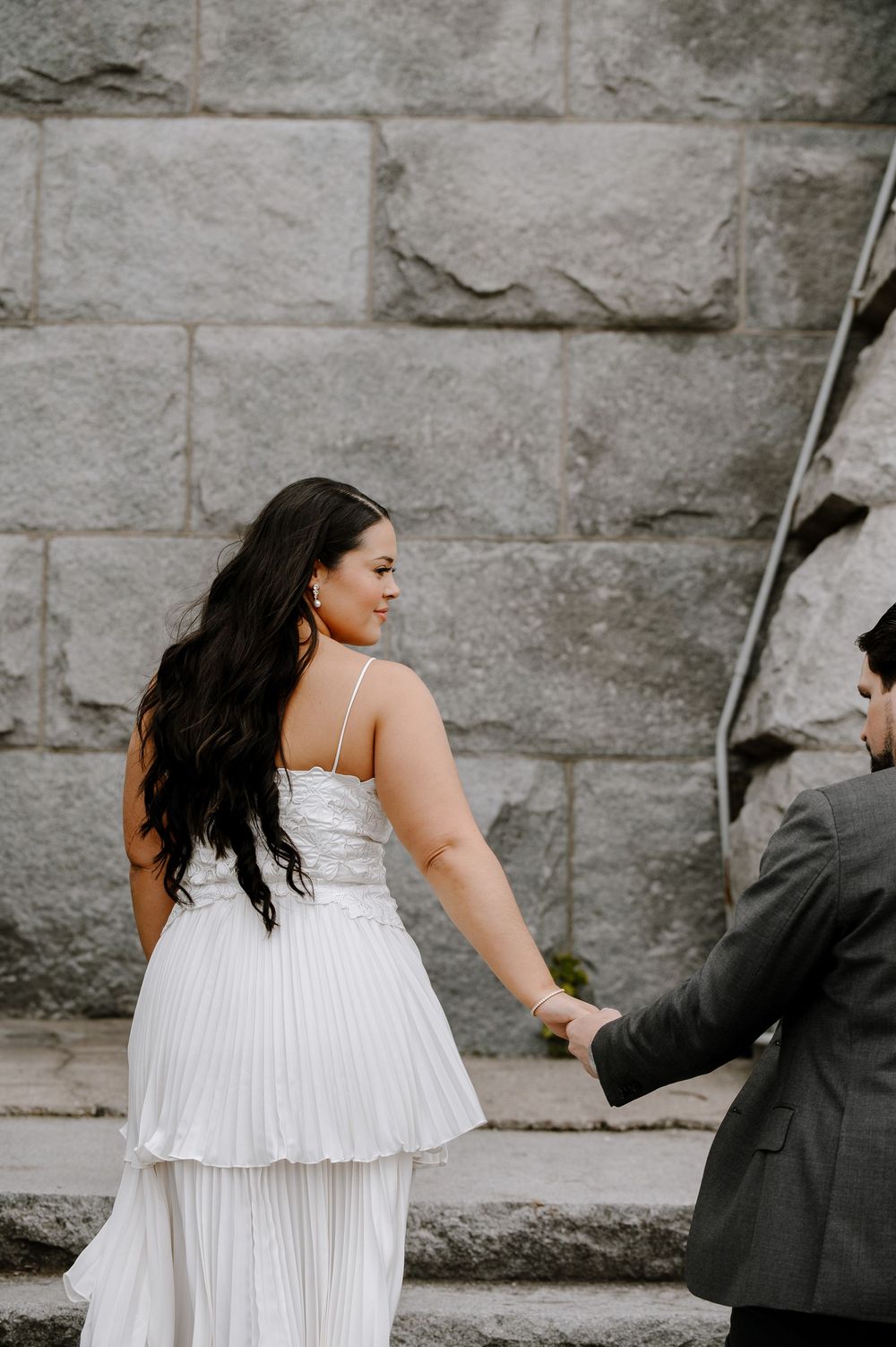 A person in a white pleated dress walks along a stone wall while holding hands with someone in a dark suit.