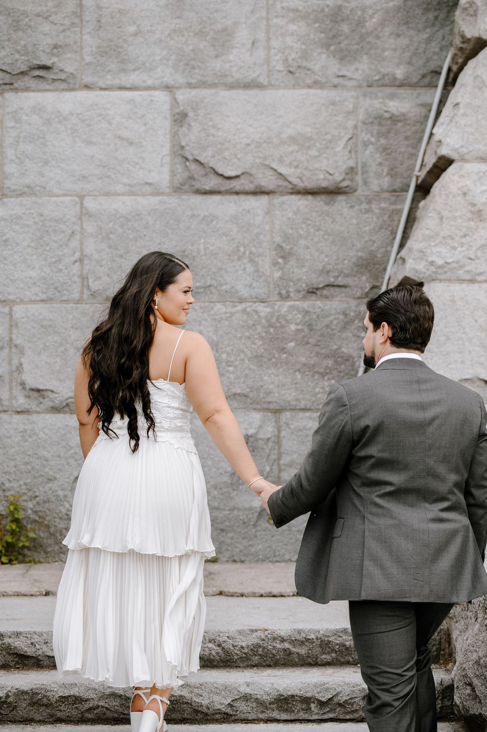A couple in wedding attire walking up stone steps together with a grey stone wall in the background.