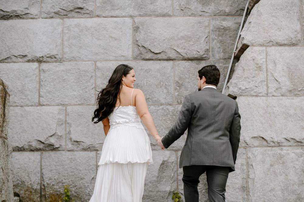 A couple in wedding attire stands against a stone wall, with the bride wearing a flowing white dress and groom in grey suit.