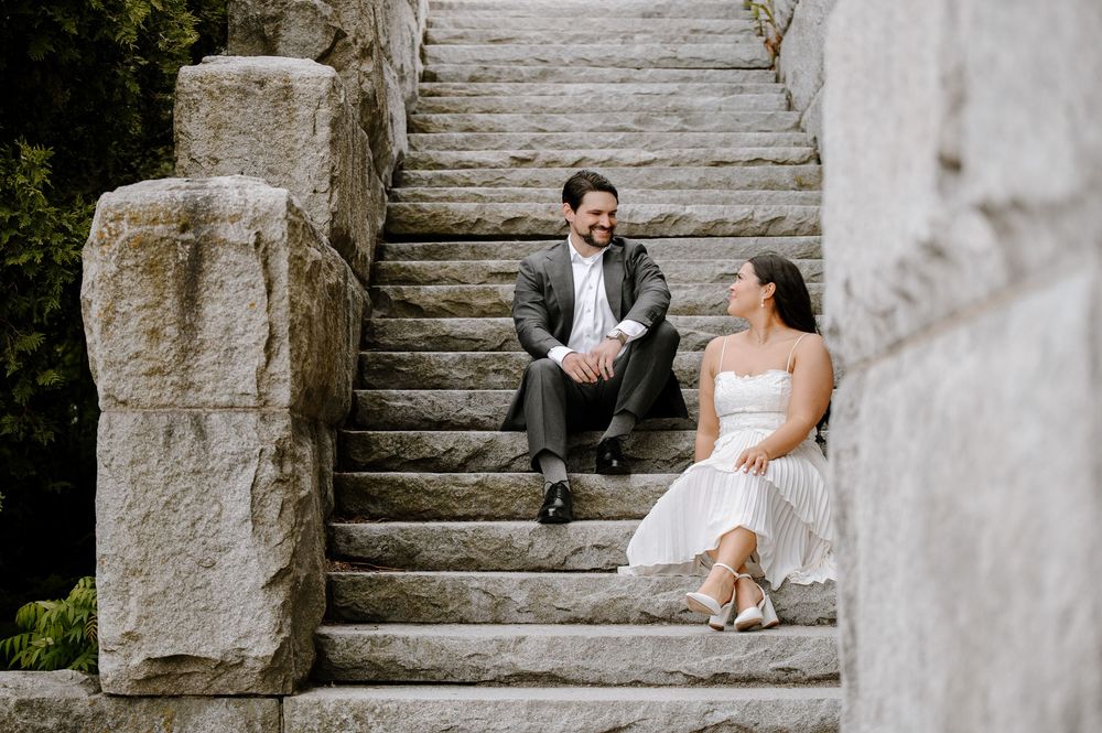 A couple in wedding attire sits together lovingly on stone steps in an elegant outdoor setting during a photoshoot.