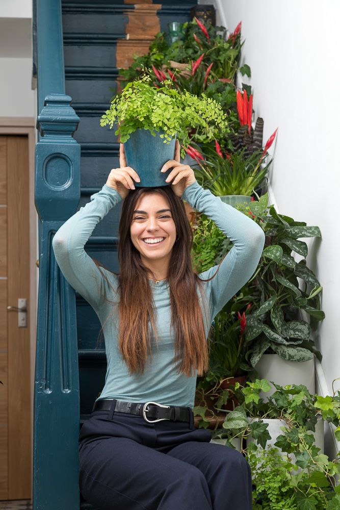 A person in a light blue sweater sits outdoors near plants and greenery on a porch.