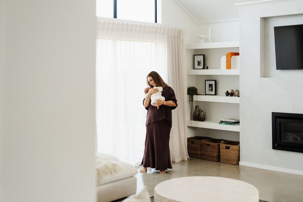 A person in a brown dress stands in a bright minimalist living room next to built-in shelving and large windows.