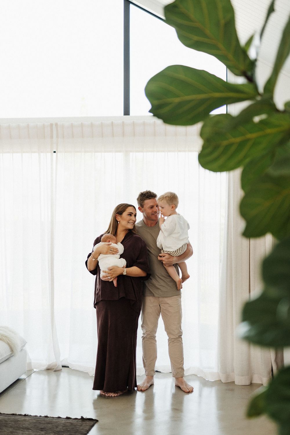 Light-filled studio maternity session showing a tender family moment against sheer white curtains and green plants.
