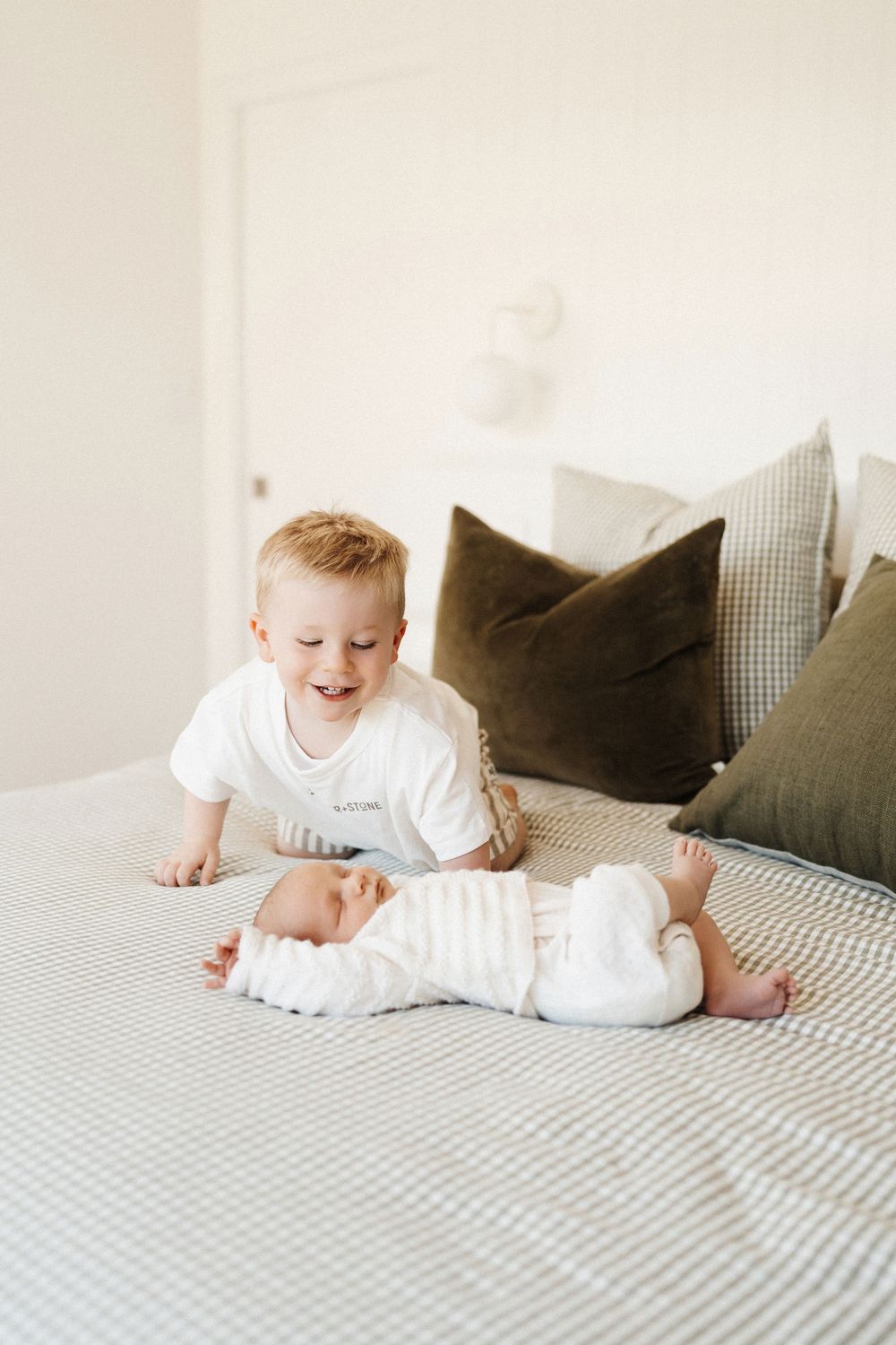 Two children on a gray checkered bed with throw pillows, one in white clothing smiling while playing together.
