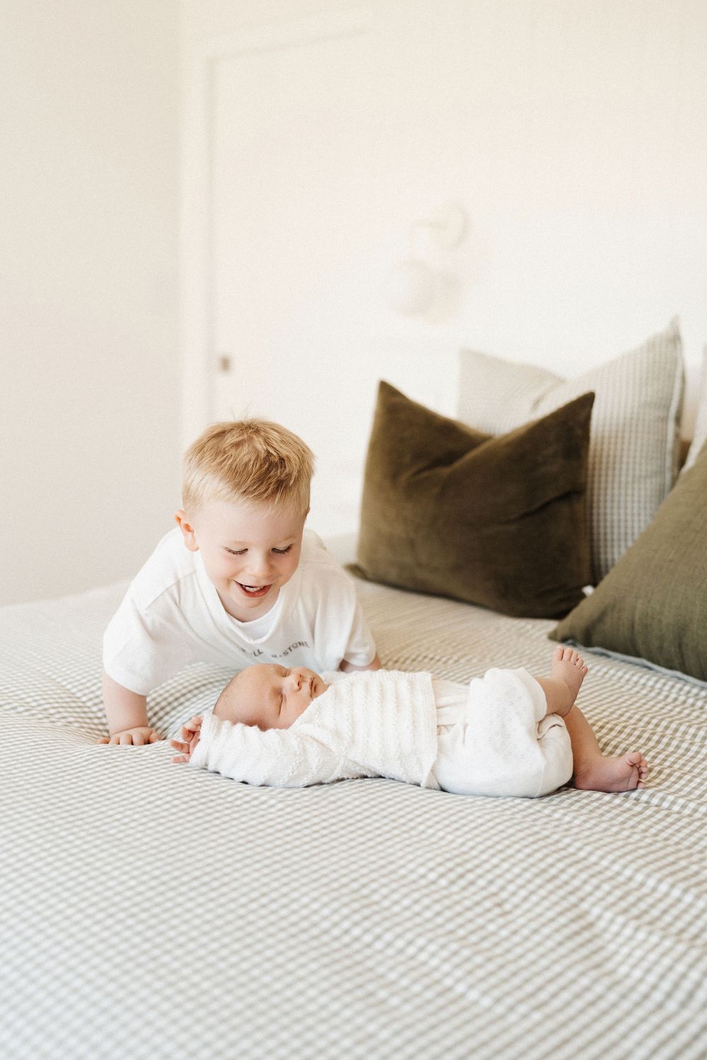 Two young children in white clothes on a gray and white bed with olive green throw pillows in the background.