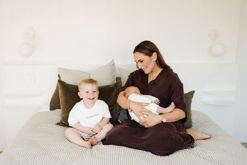 A person in brown dress sits on gray bed with two young children in white outfits.