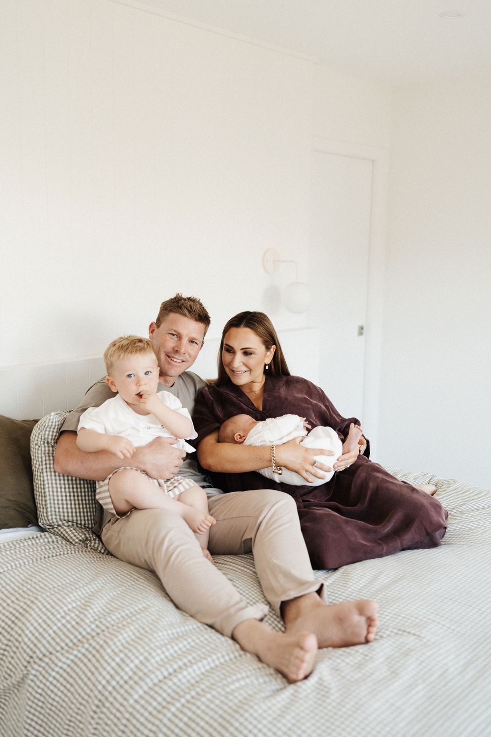 A family relaxes together on a grey bedspread in a minimalist white room while holding their young children.