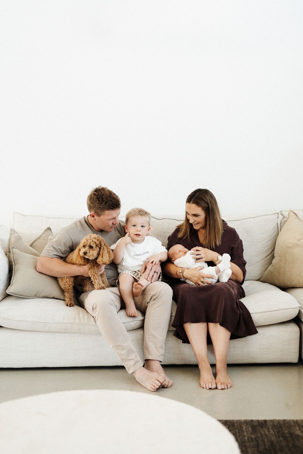 A family lounges together in cozy embrace on a light gray sofa, showcasing tender moments of connection and affection.