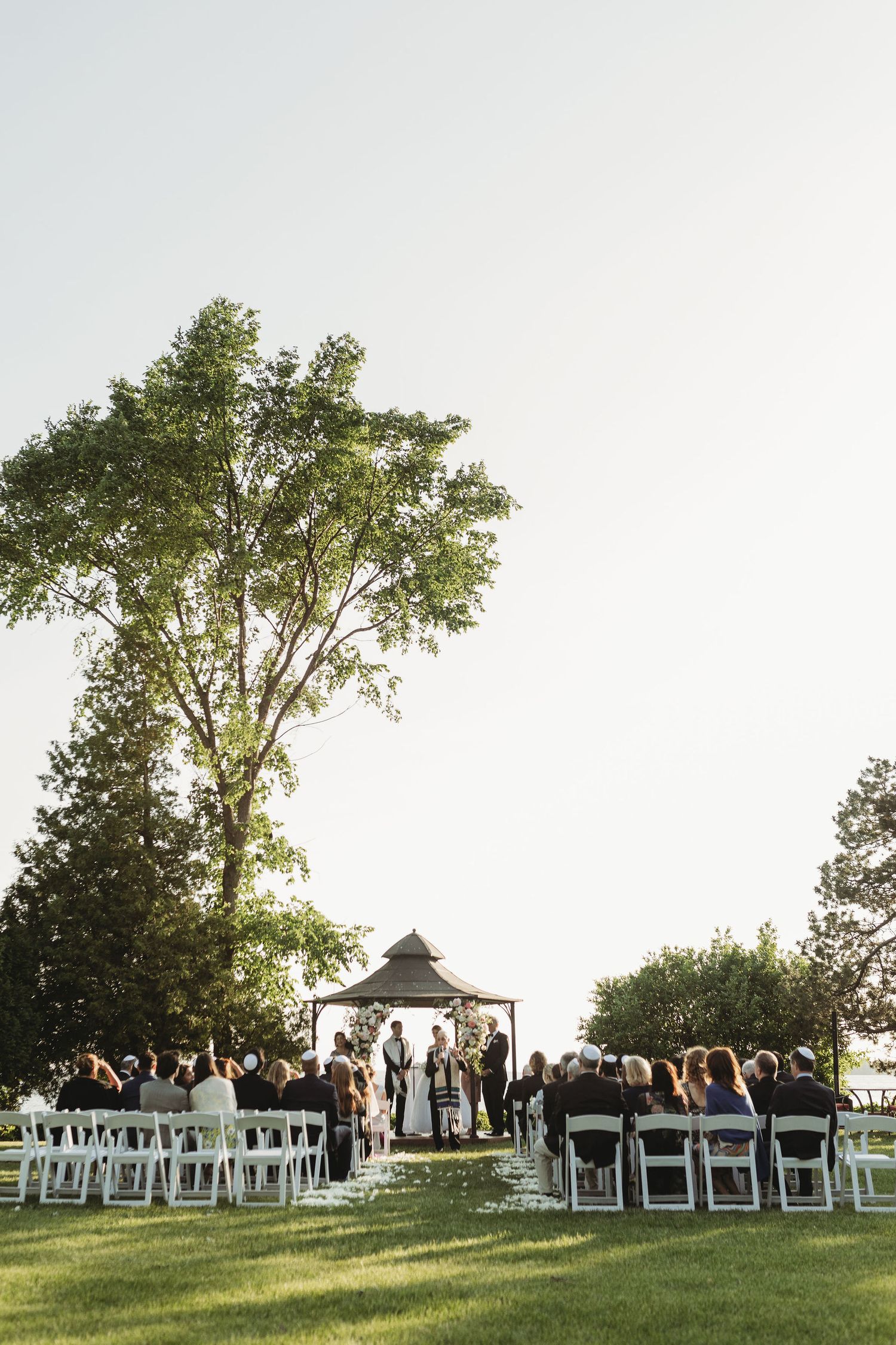 An outdoor wedding ceremony setup with white chairs and gazebos on a hillside under tall trees at sunset.