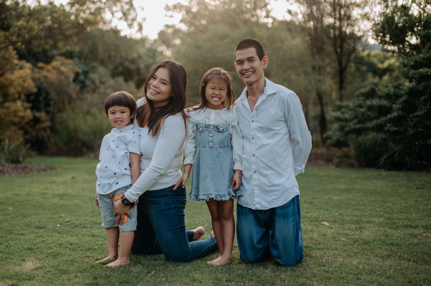 A happy family of four poses together outdoors on a grassy lawn during golden hour with trees in the background.